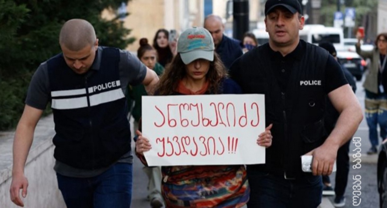 Полицейские ведут активистку, держащую плакат с надписью «Анцухелидзе бессмертен!». Фото: Леван Зазадзе / Pirveli https://tvpirveli.ge/ka/siaxleebi/sazogadoeba/103394-aqtivisti-plakatit-antsukhelidze-ukvdavia-5000-larit-daajarimes